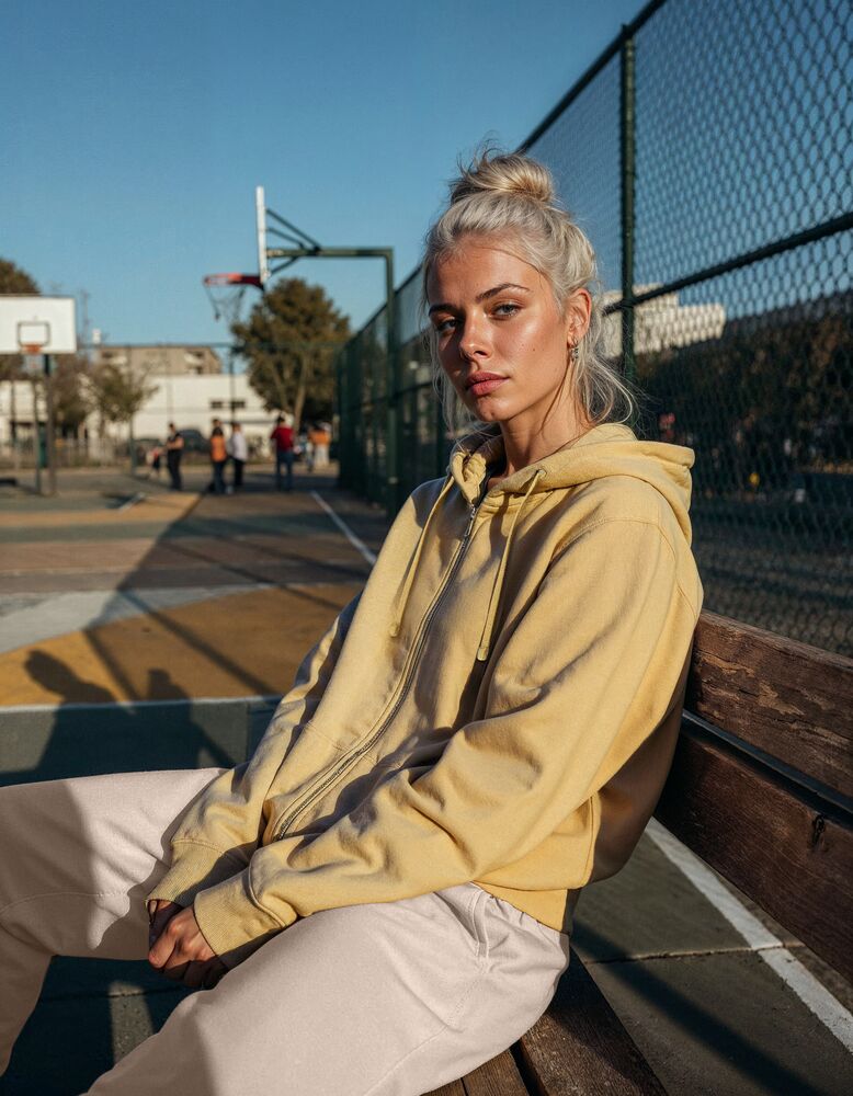 Person i gul hoodie sitter på en bänk vid en basketplan i kvällssol - symboliserar stil och komfort i Jobbutrustnings sweatshirts.