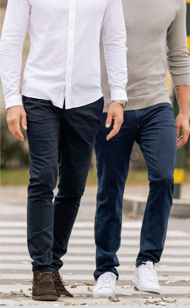 Två personer går över ett övergångsställe. Den ena bär vit skjorta, mörkblå byxor och bruna skor. Den andra har grå långärmad tröja, mörkblå jeans och vita sneakers. Bakgrunden visar en stadsmiljö med grönska och en gulsvart stolpe.