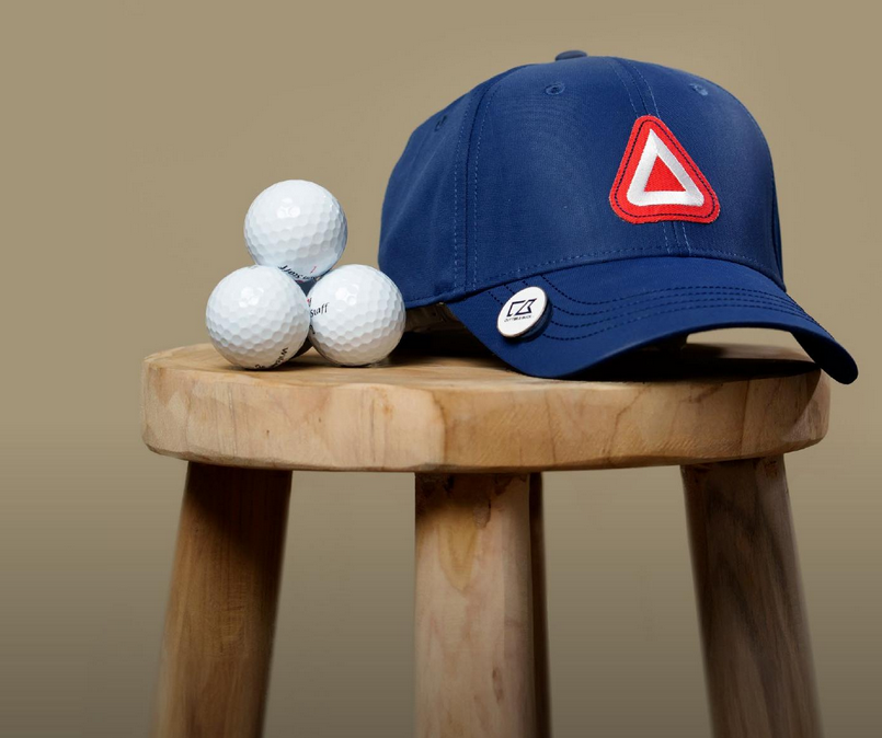Mörkblå golfkeps med rött och vitt triangelformat märke på framsidan, placerad på en rund träpall bredvid tre vita golfbollar. Perfekt keps för golf och sport, justerbar passform och stilren design.