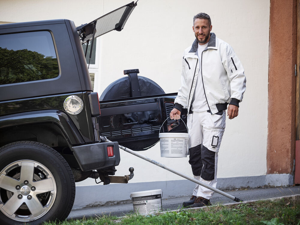 Man i målarställ står vid SUV med öppet bagage, håller färghink och verktyg – redo för utomhusarbete.