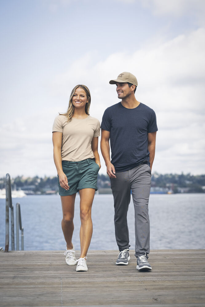 Två personer promenerar på en träbrygga vid vatten. Den ena bär beige t-shirt, gröna shorts och vita sneakers. Den andra har marinblå t-shirt, grå byxor, grå sneakers och beige keps. Bakgrunden visar vatten, träd och molnig himmel.